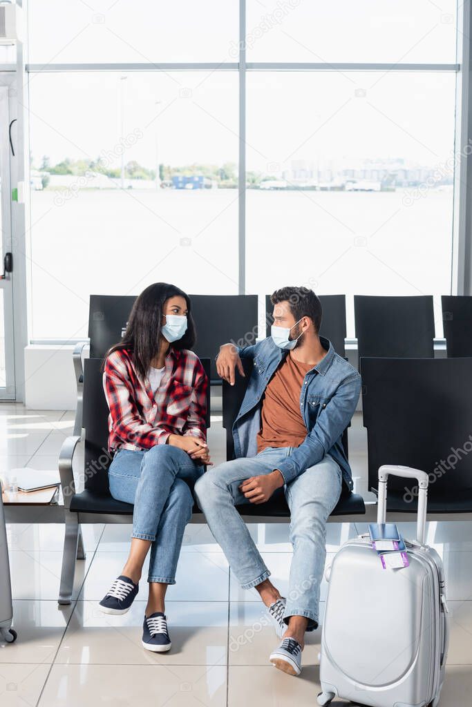 interracial couple in medical masks sitting and looking at each other near luggage in departure lounge 