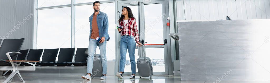 happy multicultural couple walking with luggage and passports in airport, banner