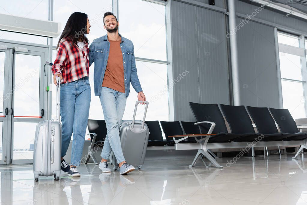 cheerful multicultural couple walking with luggage in airport 