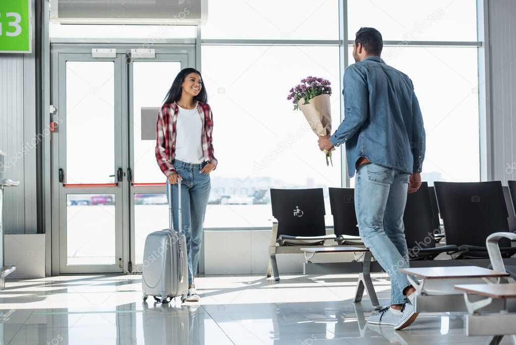 man holding wrapped flowers while meeting cheerful african american girlfriend in airport