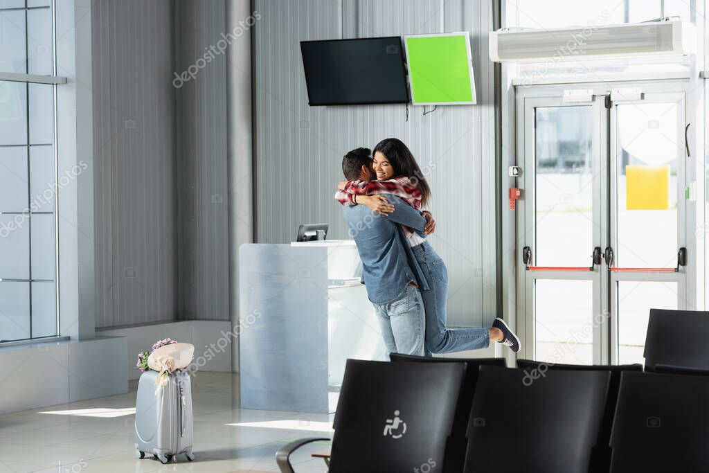 man holding in arms cheerful african american woman while meeting near luggage in airport 