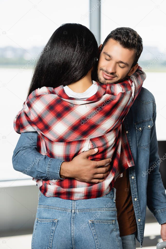 cheerful man smiling while hugging african american woman in airport 