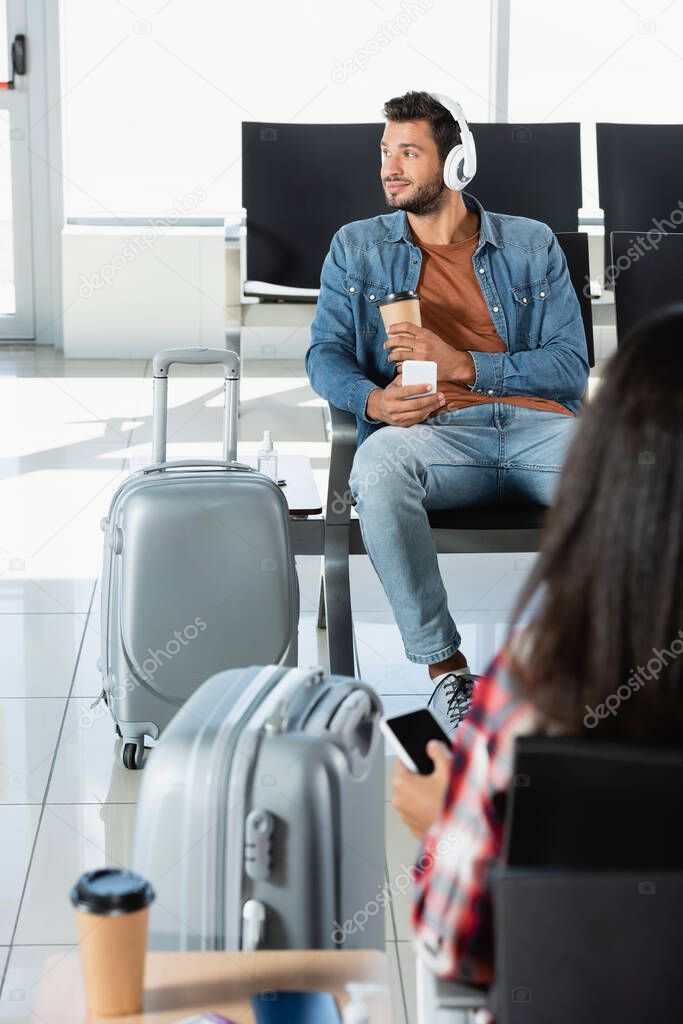 smiling man in headphones holding paper cup and smartphone near luggage and woman on blurred foreground 