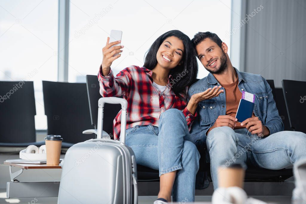 happy african american woman pointing with hand while taking selfie with bearded man holding passport in airport