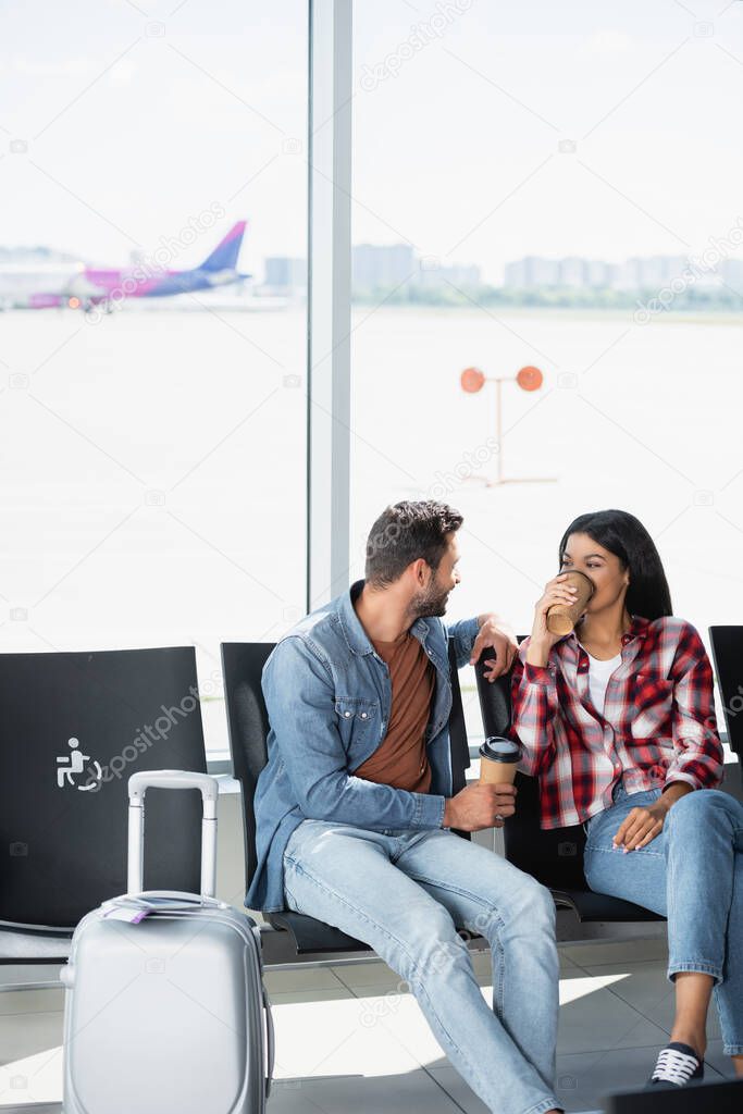 african american woman drinking coffee to go near bearded man in departure lounge of airport 