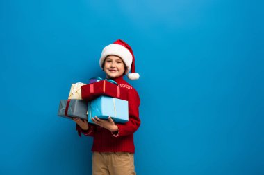 Noel Baba şapkalı ve süveterli mutlu çocuk mavi üzerinde hediyeler tutuyor.