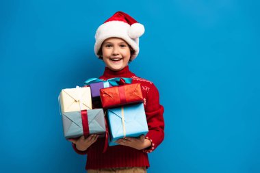 Noel Baba şapkalı ve süveterli neşeli çocuk mavi üzerinde hediyeler tutuyor.