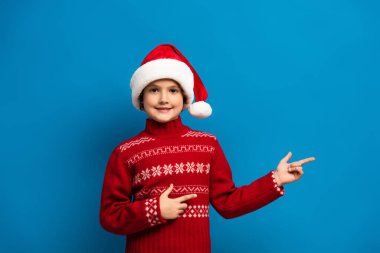 Noel Baba şapkalı neşeli çocuk ve kırmızı süveter parmağıyla maviyi gösteriyor.