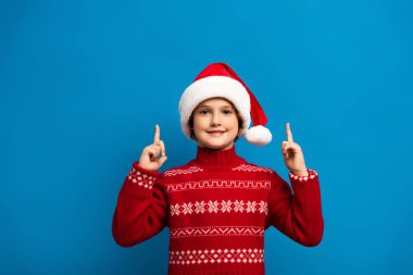 Noel Baba şapkalı ve kırmızı kazaklı neşeli çocuk mavi kameraya bakarken parmaklarıyla işaret ediyor.