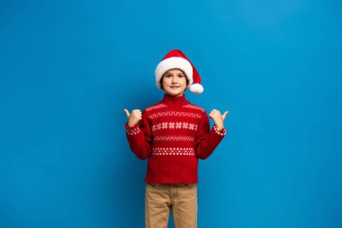 Noel Baba şapkalı ve kırmızı kazaklı neşeli çocuk mavi kameraya bakarken başparmağını kaldırıyor.