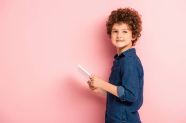 Memnun ve kıvırcık çocuk dijital tablet tutuyor ve pembe kameraya bakıyor. 
