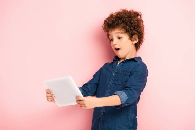 Şaşırmış ve kıvırcık çocuk pembe üzerinde dijital tablet kullanıyor 