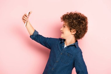 Kotlu tişörtlü kıvırcık çocuk pembe telefondan selfie çekiyor.