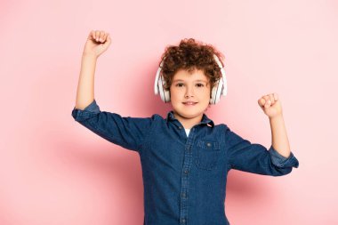 Heyecanlı ve kıvırcık çocuk, yumruklarını sıkmış, pembe kulaklıkla müzik dinliyor. 