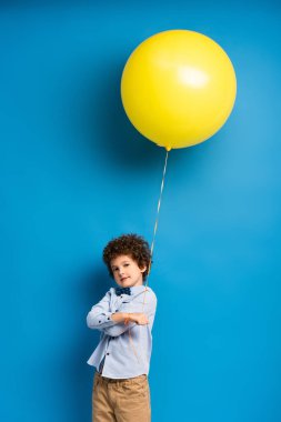 Kıvırcık tişörtlü ve papyonlu çocuk mavi üzerinde sarı balon tutuyor.