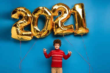 Kırmızı kazaklı kıvırcık çocuk mavi üzerinde 2021 numara olan altın balonların yanında baş parmağını gösteriyor.