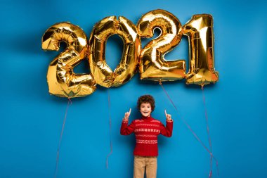 Kırmızı kazaklı heyecanlı çocuk mavi üzerinde 2021 numara olan altın balonları işaret ediyor.