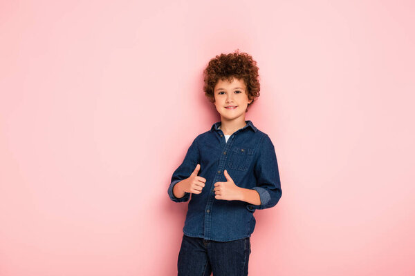 joyful and curly boy showing thumbs up on pink 