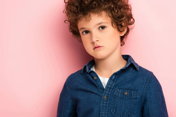 serious and curly boy in denim shirt looking at camera on pink 