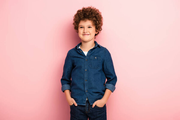 pleased and curly boy in denim shirt standing with hands in pockets on pink 