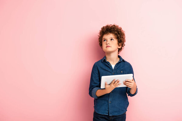 curly boy in denim shirt looking up and holding digital tablet on pink 