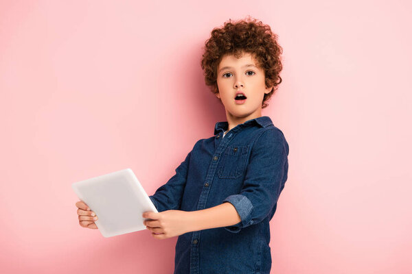 shocked and curly kid holding digital tablet on pink 