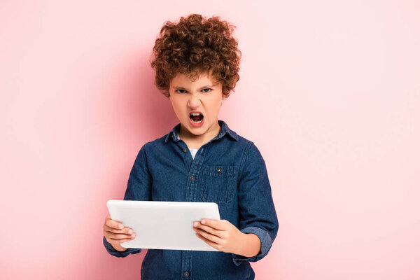 angry and curly kid screaming and holding digital tablet on pink 