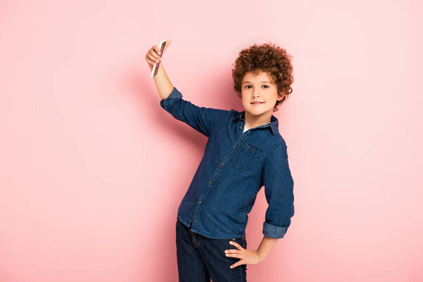 curly kid in denim shirt taking selfie on smartphone while standing with hand on hip on pink