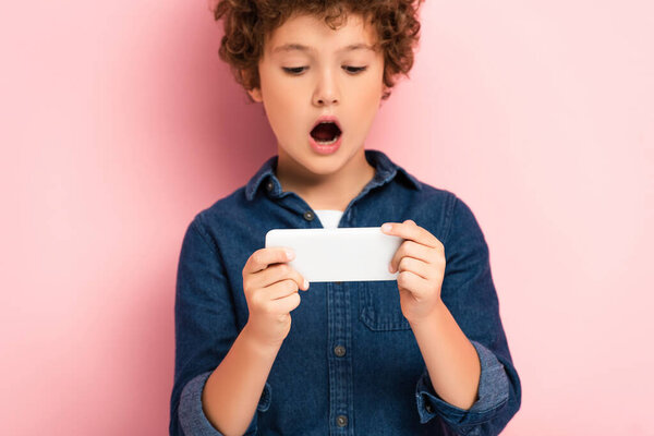 selective focus of surprised boy in denim shirt using smartphone on pink