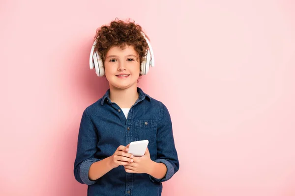 Joyful Curly Boy Listening Music Wireless Headphones Holding Smartphone Pink — Stock Photo, Image