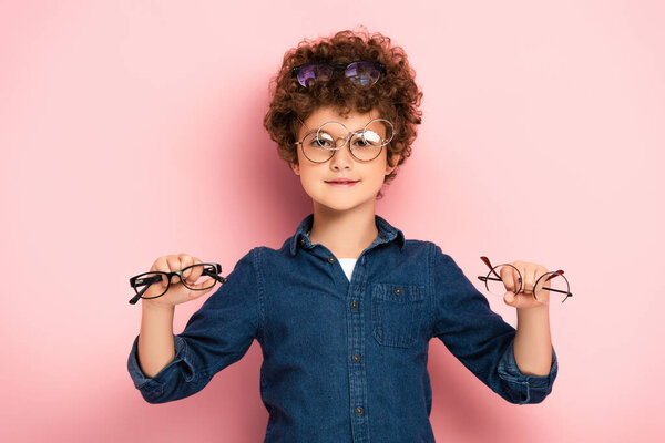 joyful boy holding bunch of eyeglasses while looking at camera on pink  