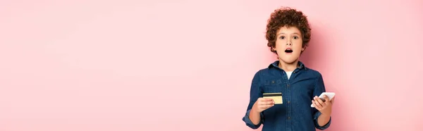 ショックを受けた少年がスマートフォンやクレジットカードを持ちながらピンクでオンラインショッピングをするというパノラマのコンセプト — ストック写真