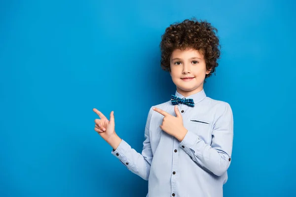 curly kid in shirt and bow tie pointing with fingers on blue