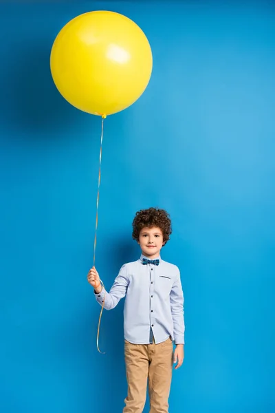 happy boy in shirt and bow tie holding big yellow balloon on blue