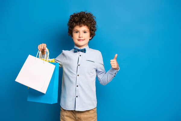 joyful and curly boy holding shopping bags and showing thumb up on blue 
