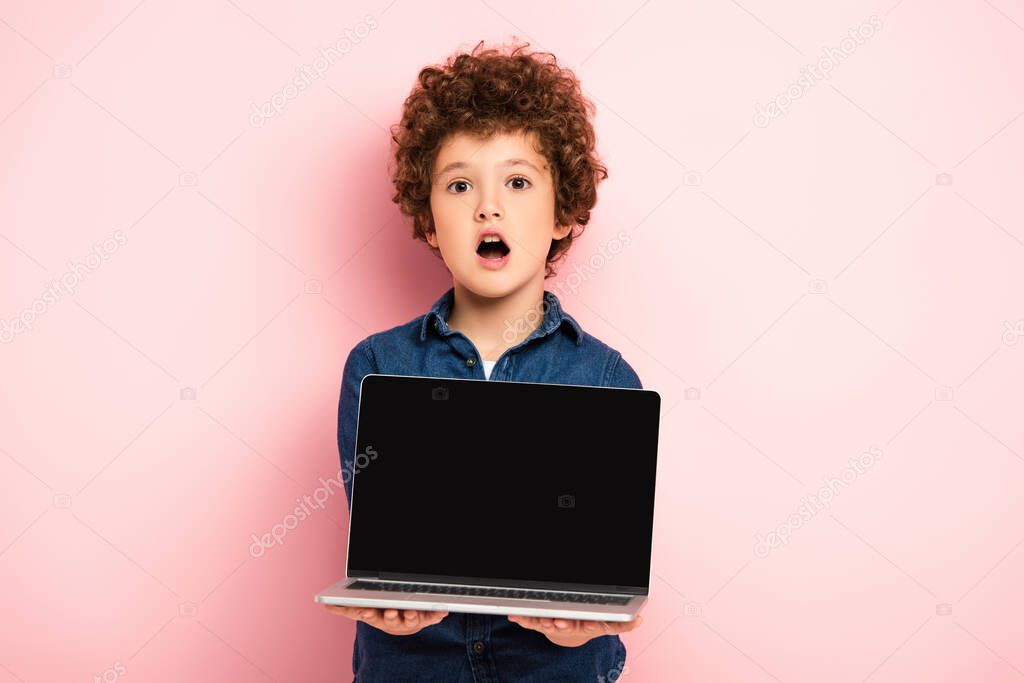 shocked and curly boy holding laptop with blank screen on pink 