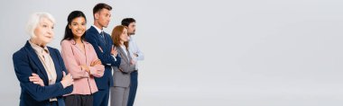 African american businesswoman smiling at camera near colleagues with crossed arms on blurred background isolated on grey, banner 