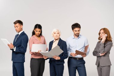Multicultural businesspeople using gadgets, holding paper folders and talking on smartphone isolated on grey
