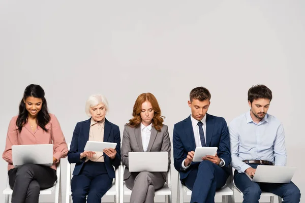 Empresarios Multiculturales Usando Dispositivos Mientras Están Sentados Sillas Aisladas Gris —  Fotos de Stock