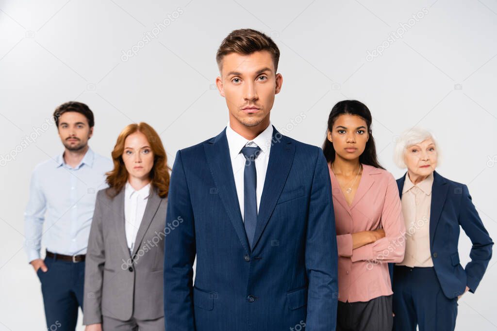 Businessman in suit looking at camera near multicultural colleagues on blurred background isolated on grey