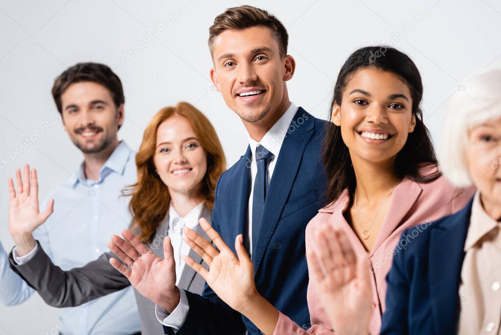 Cheerful businessman waving hand near multicultural colleagues on blurred background isolated on grey