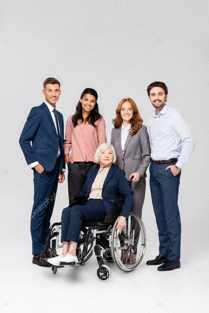 Positive multiethnic businesspeople looking at camera near senior colleague in wheelchair on grey background 