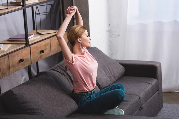 Вид сбоку привлекательной молодой женщины с поднятыми руками, сидящей дома на диване — стоковое фото