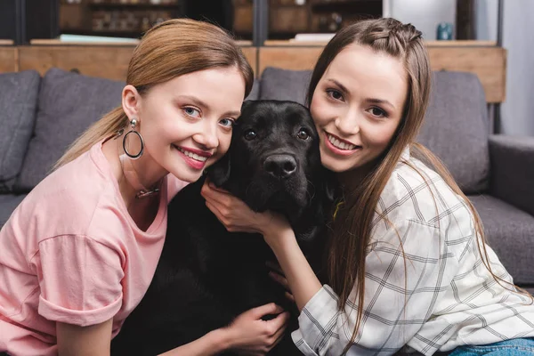 Smiling young female friends embracing black retriever at home — Stock Photo