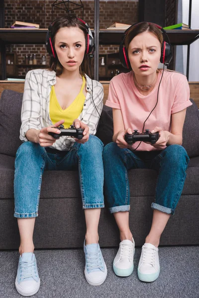 Amis féminins concentrés dans des écouteurs avec joysticks dans les mains jouer à un jeu vidéo — Photo de stock