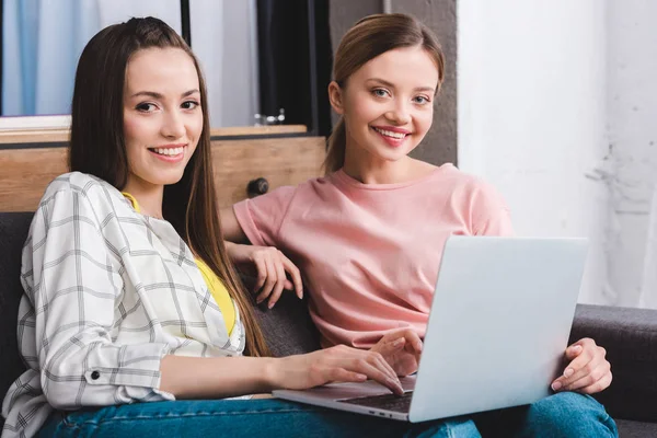 Smiling young female friends with laptop sitting on sofa at home — Stock Photo