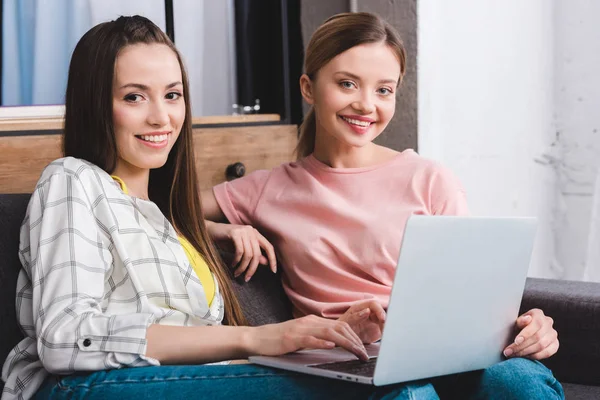 Sonrientes amigas jóvenes con portátil sentado en el sofá en casa - foto de stock