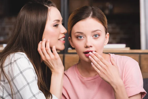 Vista lateral de mujer joven susurrando secreto a amiga sorprendida - foto de stock