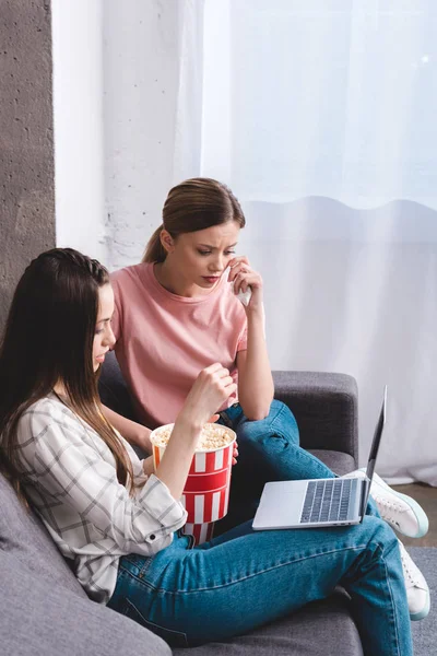 Crying young woman watching movie with female friend eating popcorn and holding laptop on knees at home — Stock Photo