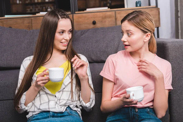 Dos amigas jóvenes con tazas de café sentadas en el sofá en casa - foto de stock